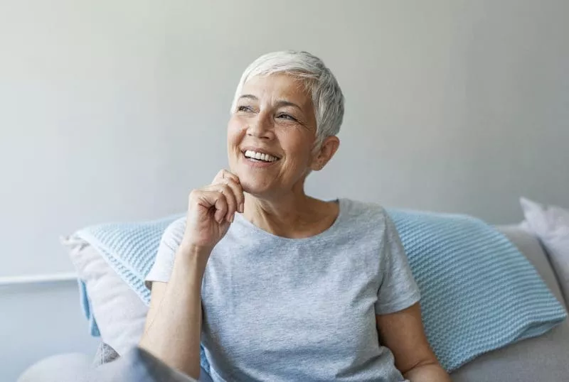 Smiling woman with short gray hair sitting on a sofa, resting her chin on her hand and looking thoughtfully to the side.