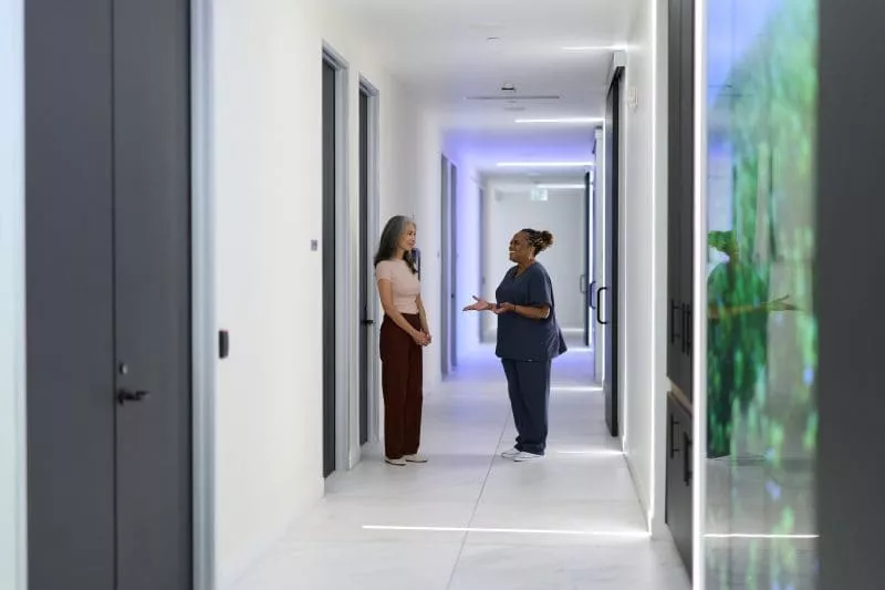 Two women talking in a bright, modern office hallway with glass doors and soft lighting.