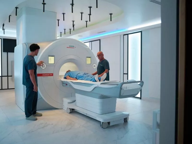 Patient on a hospital bed being positioned into an MRI scanner by two clinicians in a modern imaging suite.
