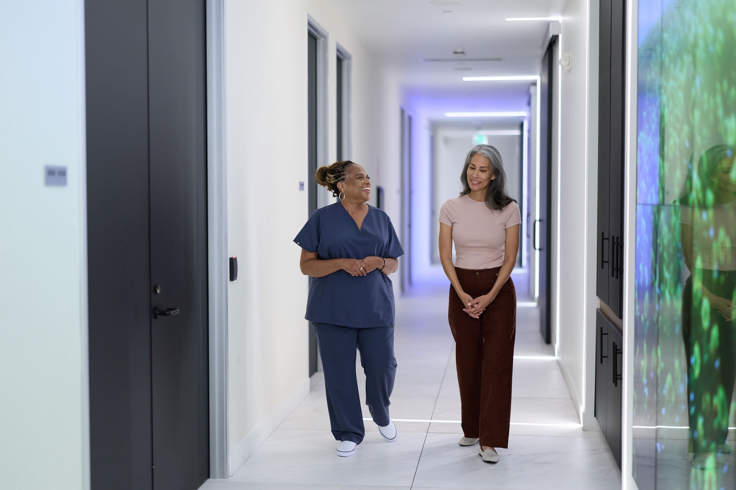 Two women walking and talking in a bright, modern hallway, one wearing scrubs and the other in business-casual clothing.