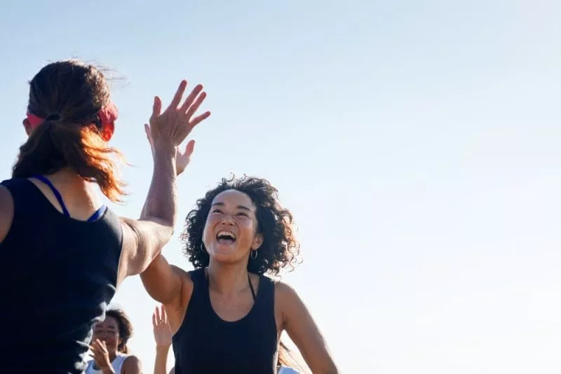 Two women high-fiving and smiling outdoors during a group fitness session.