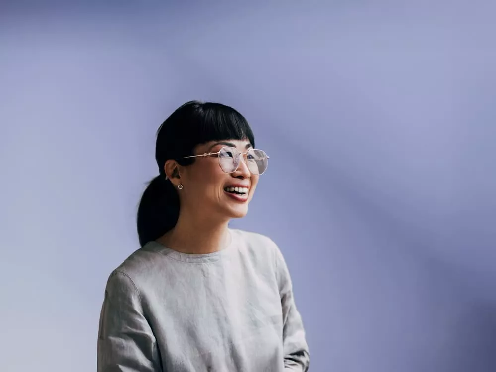Smiling woman with glasses and a low ponytail looking to the side against a soft blue background.