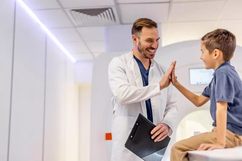 Healthcare professional in a white coat high-fiving a smiling child sitting on an MRI scanner bed.