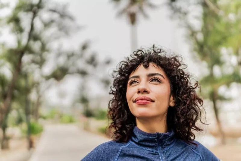 Woman with curly hair wearing a blue jacket, looking upward while standing on a tree-lined path outdoors.