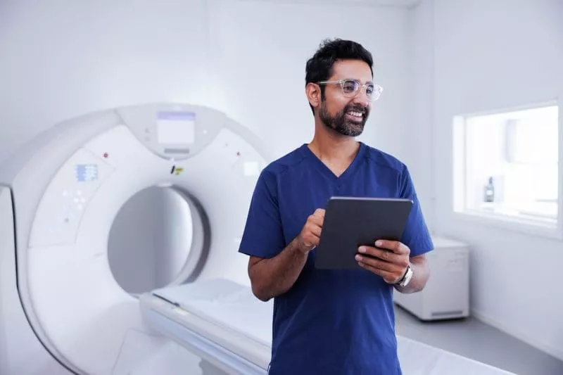 Clinician holding a tablet beside a CT scanner in a medical imaging room.