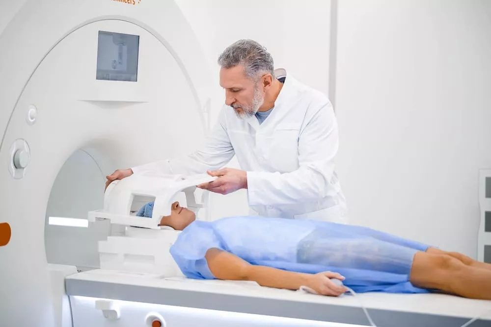 Clinician adjusting a head coil on a patient lying on an MRI scanner table in a bright clinical room.