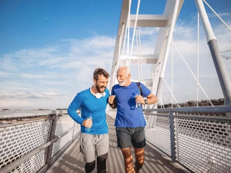 Two men jogging together across a pedestrian bridge, chatting and holding water bottles under a blue sky.