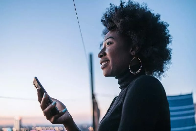 Woman smiling while looking at her phone outdoors at dusk.