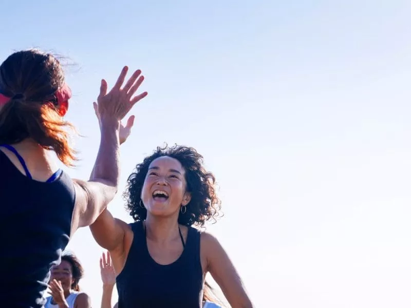 Two women in sportswear high-fiving outdoors, smiling during a group fitness session.
