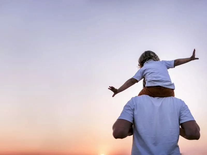 Adult carrying a child on their shoulders with arms outstretched at sunset.