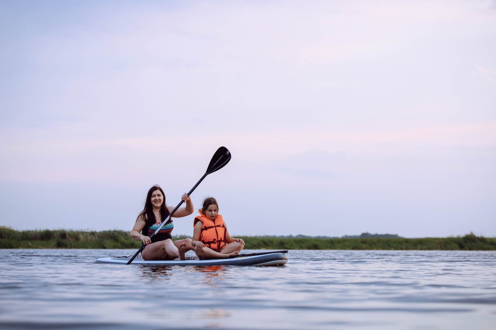 Woman paddleboarding with a young child wearing a life jacket on calm water at dusk.