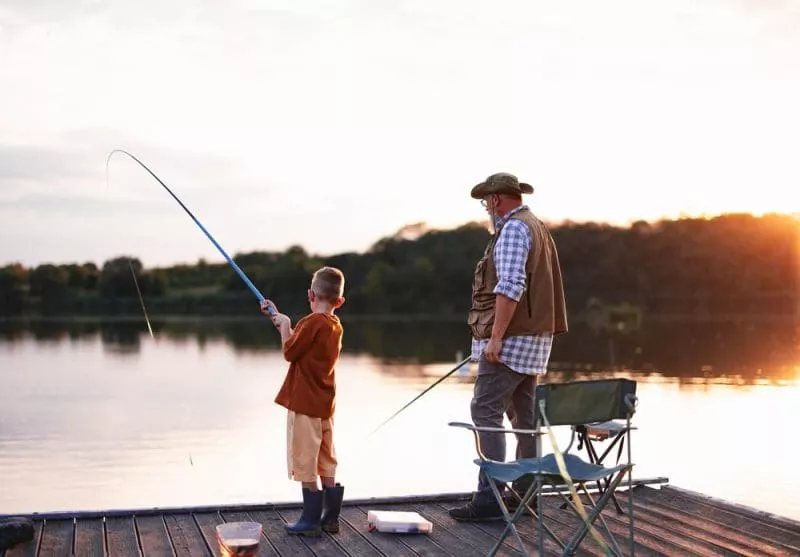 An older man and a young boy fishing together from a wooden dock at a calm lake during sunset.