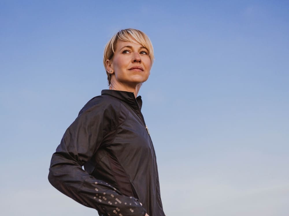 Woman with short blonde hair in a black jacket standing outdoors, looking into the distance against a clear blue sky.