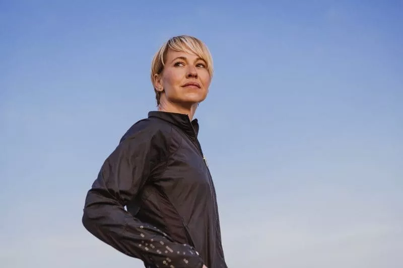 Person stretching with arms raised overhead by the water, wearing a hoodie and gloves.
