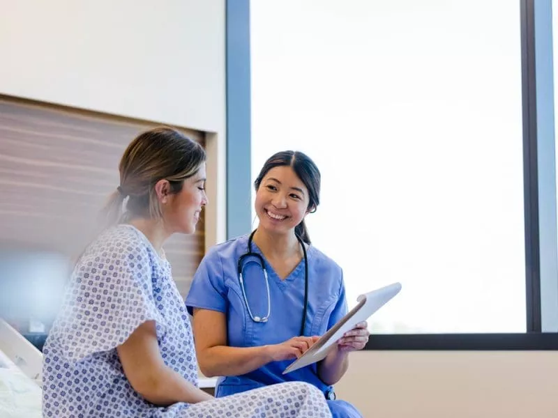 Nurse in blue scrubs smiling while reviewing a tablet with a patient sitting on a hospital bed.