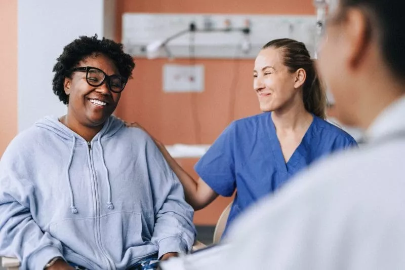 Smiling patient in a hoodie sitting with two healthcare professionals, one gently resting a hand on her shoulder in a hospital room.