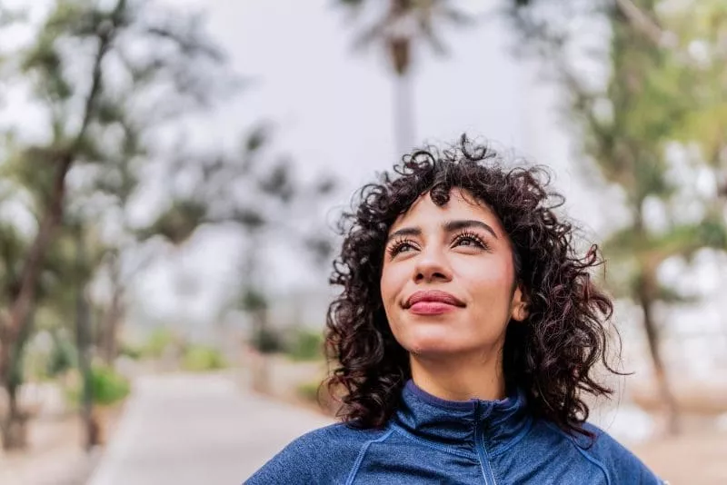 Woman with curly hair wearing a blue jacket, looking upward while standing on a tree-lined path outdoors.
