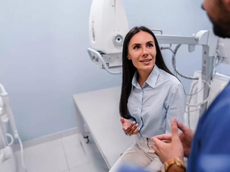 Woman seated in a medical imaging room talking with a clinician, with scanning equipment visible behind her.
