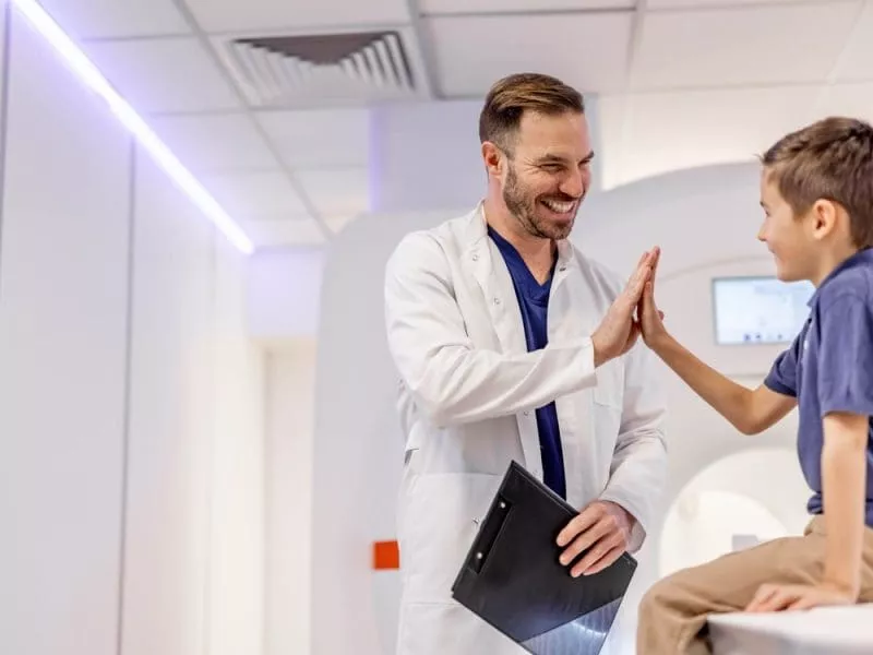 Healthcare professional in a white coat high-fiving a smiling child sitting on an MRI scanner bed.