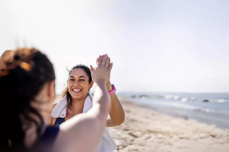 Two people high-fiving on the beach after a workout, smiling with the ocean in the background.