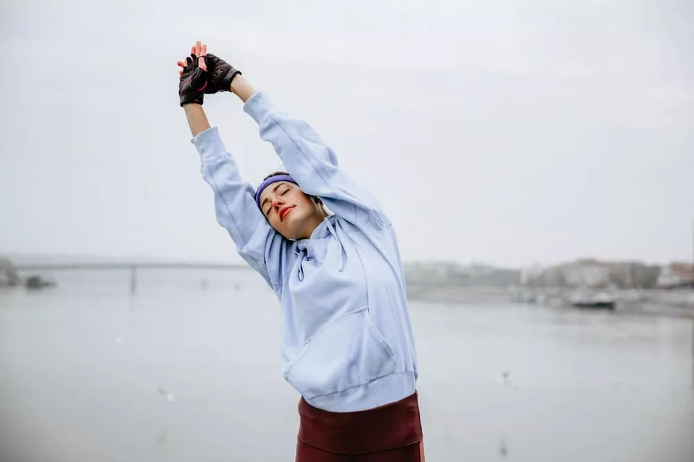 Person stretching with arms raised overhead by the water, wearing a hoodie and gloves.