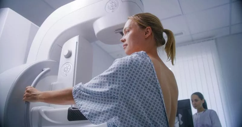 Patient positioning herself at a mammography machine while a technician stands nearby in the background.