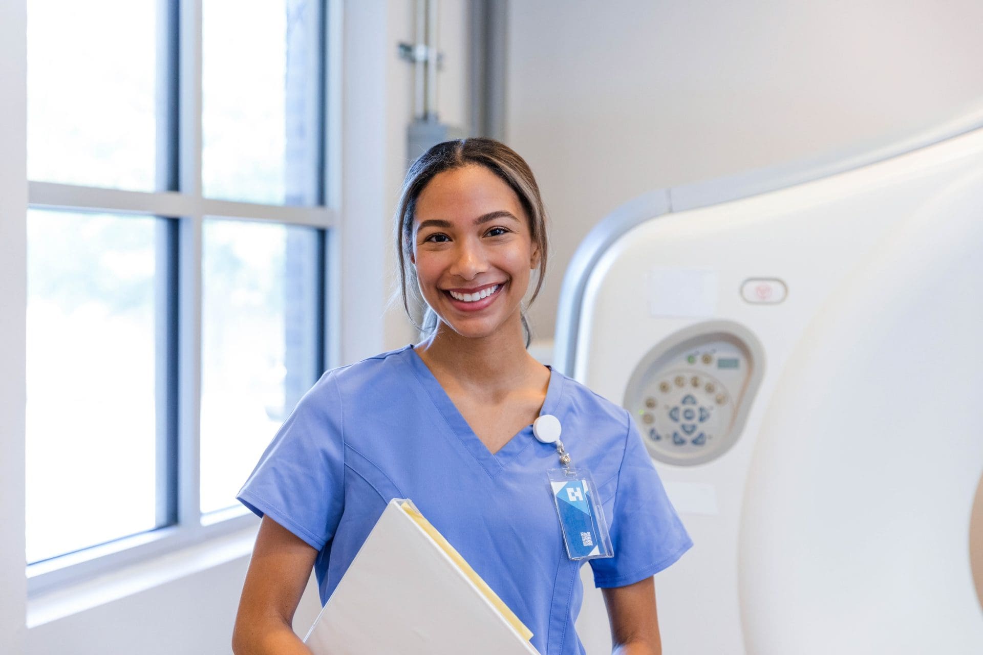Smiling healthcare professional in blue scrubs holding a folder, standing in a clinical imaging room.