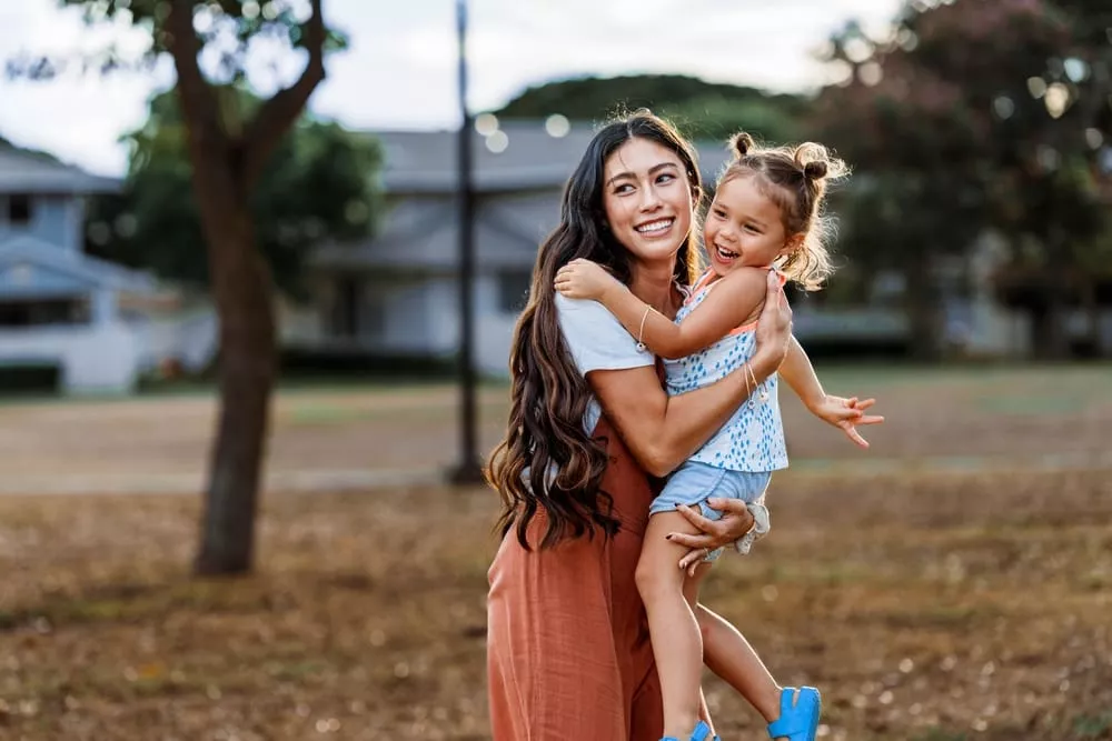 Woman holding a young child and smiling together in a park with trees and houses in the background.