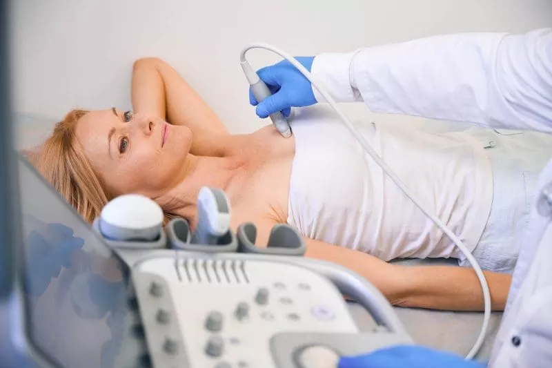 Patient lying on an exam table while a clinician performs an ultrasound scan on her chest using a handheld probe.