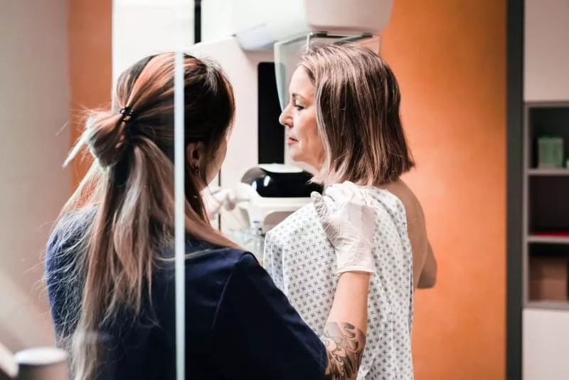 Patient standing at a mammography machine while a clinician gently positions her shoulder and upper body for the scan.