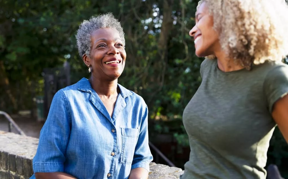 Two women smiling and talking outdoors, standing by a stone wall with trees in the background.