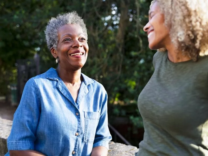 Two women smiling and talking outdoors, standing by a stone wall with trees in the background.