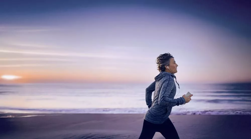 Person jogging along a beach at sunrise, wearing headphones and holding a smartphone.