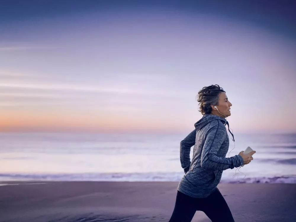 Person jogging along a beach at sunrise, wearing headphones and holding a smartphone.