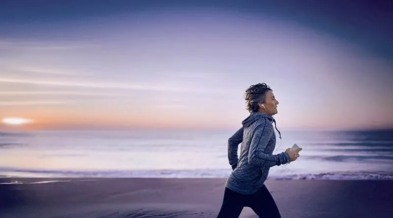 Person jogging along a beach at sunrise, wearing headphones and holding a smartphone.
