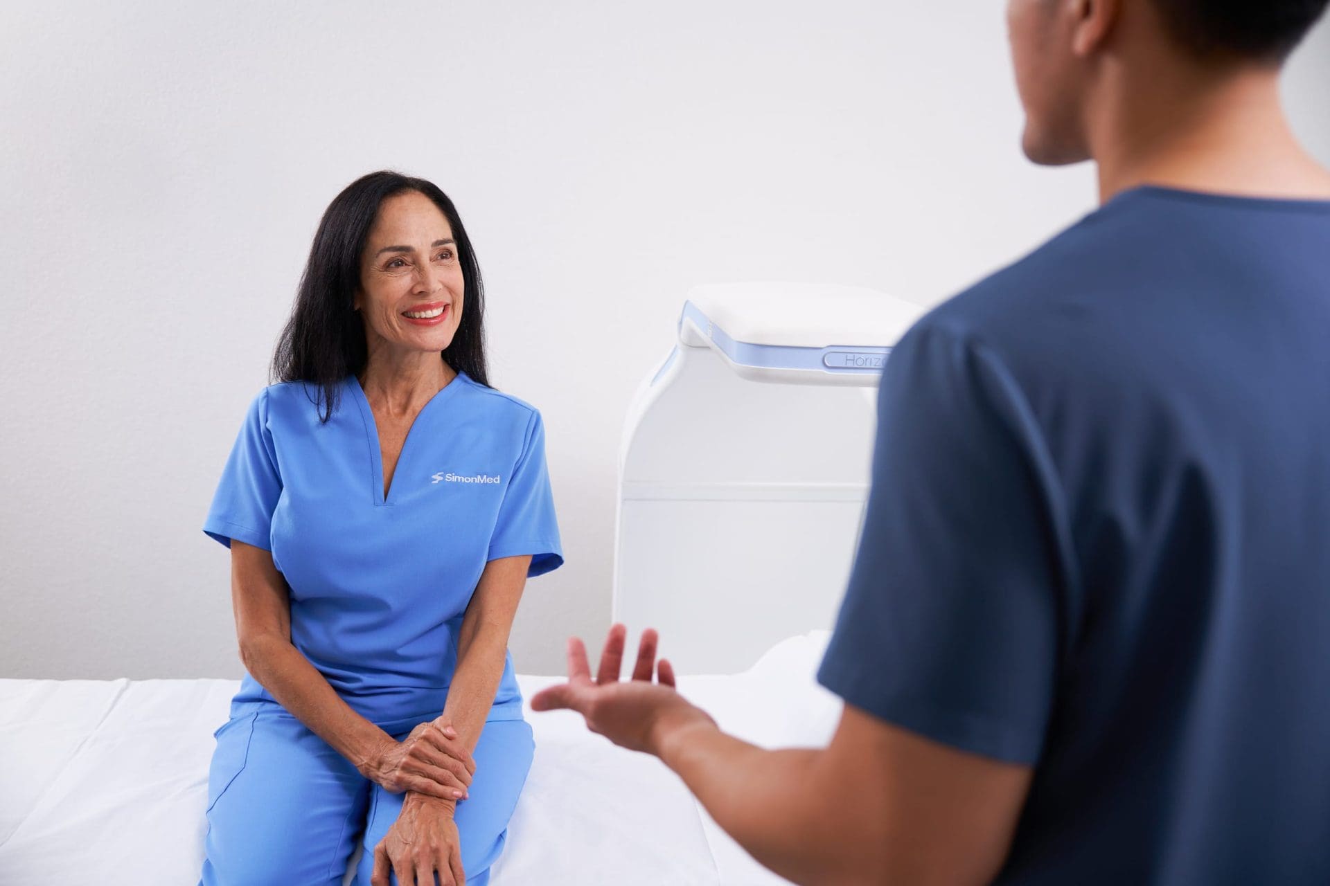 Patient sitting on an examination table, smiling while talking with a clinician in a clinical imaging room.