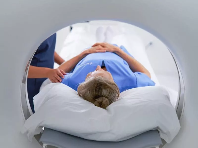 Patient lying on an MRI scanner bed, viewed from inside the scanner opening, with a clinician standing beside them.