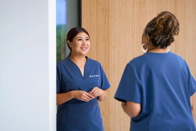Two clinic staff in navy scrubs smiling and talking in a modern reception area with a wood-panelled wall.