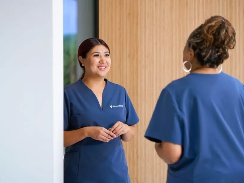 Two clinic staff in navy scrubs smiling and talking in a modern reception area with a wood-panelled wall.