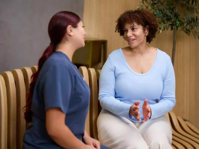A clinic staff member in navy scrubs sits talking with a woman in a light blue top on a wooden bench in a calm waiting area.