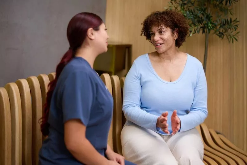 A clinic staff member in navy scrubs sits talking with a woman in a light blue top on a wooden bench in a calm waiting area.