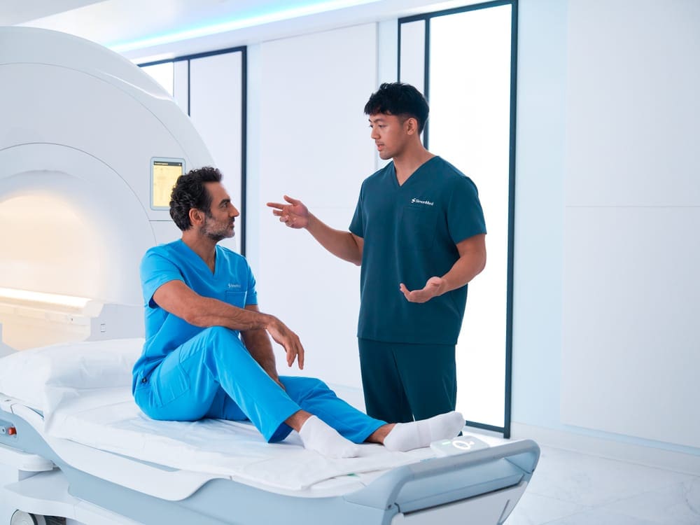 Two clinicians talking beside an MRI scanner, one seated on the scan table in a modern imaging room.