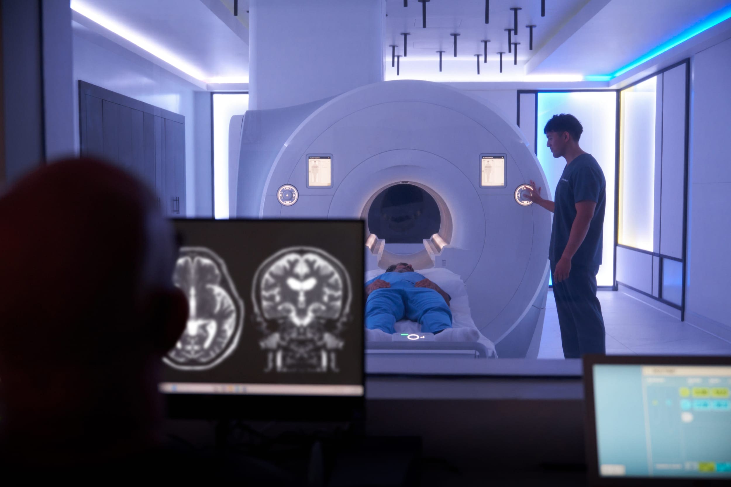 View from an MRI control room showing a patient on the scanner bed, a clinician beside the machine, and brain scan images on a monitor in the foreground.