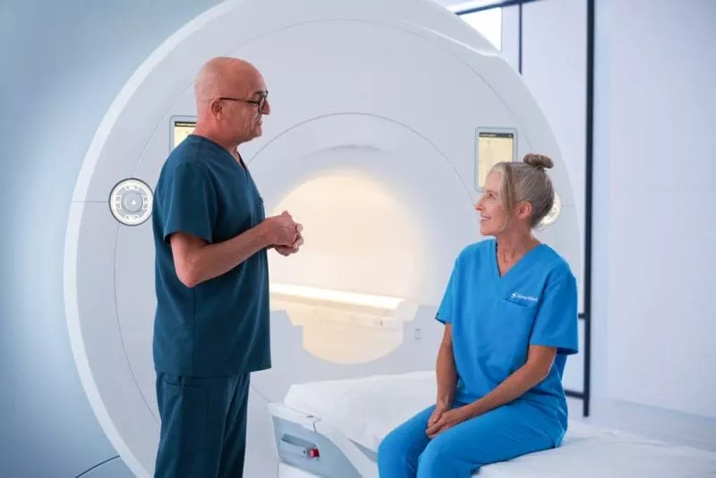 Clinician speaking with a patient seated on an MRI scanner bed in a bright imaging room.