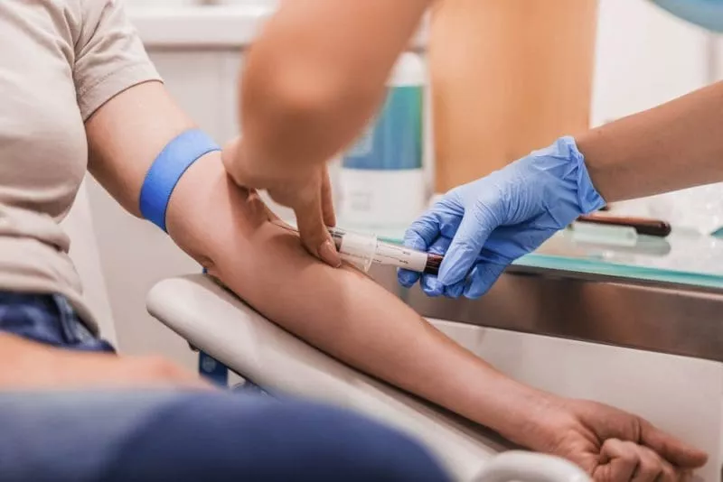 A healthcare professional wearing gloves draws blood from a patient’s arm using a syringe during a medical test.