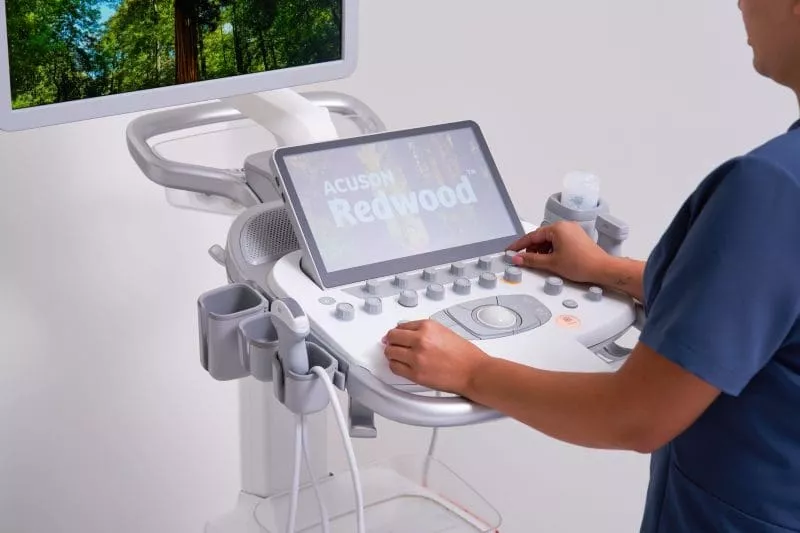 Clinician operating an ultrasound machine with a touchscreen control panel and monitor displaying the ACUSON Redwood interface.