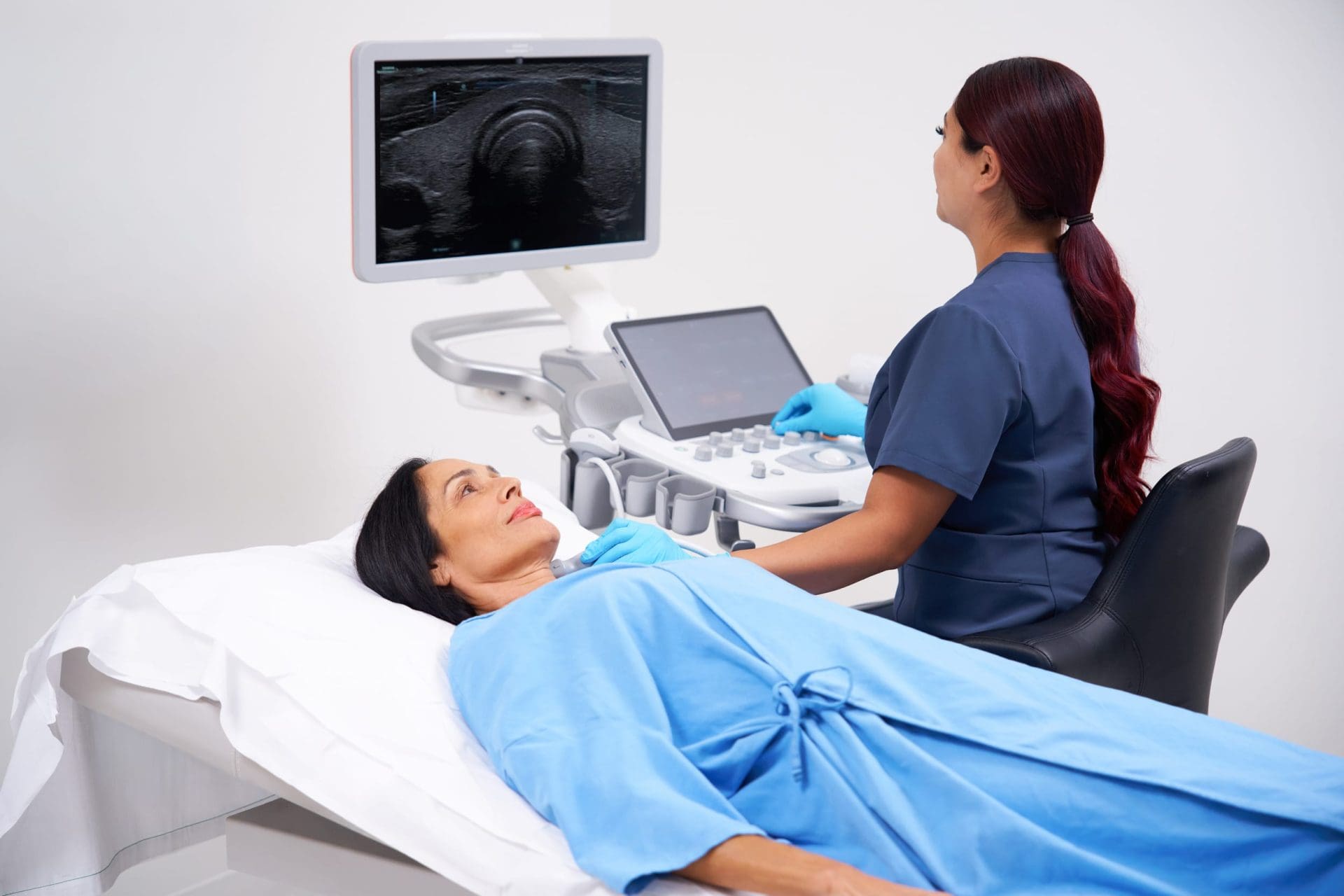 Clinician performs an ultrasound scan on a patient lying on an exam bed, with the ultrasound image displayed on a monitor.