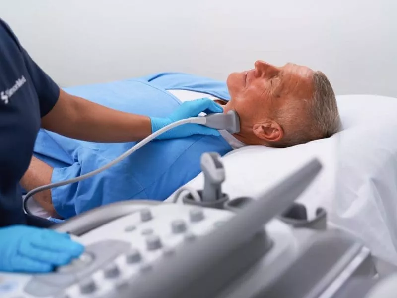 Clinician performing an ultrasound scan on an older man’s neck as he lies on an examination bed.